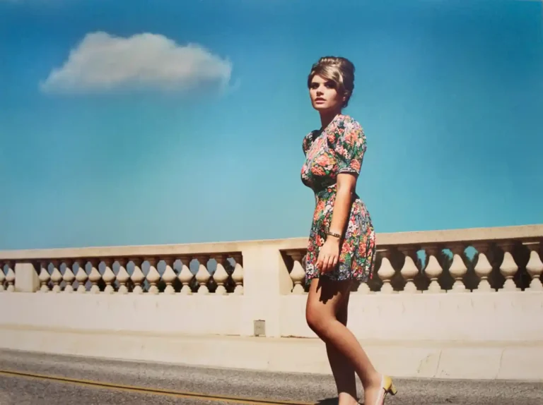 Alex Prager Art Photography poster featuring a woman in a floral dress and beehive hair, walking dramatically under a bright, cloud-filled blue sky.
