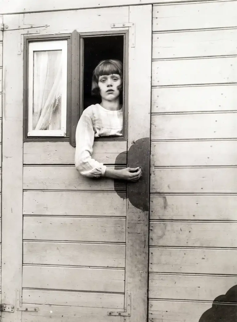 August Sander fine art photography poster featuring a young girl (Circus Girl) with a bob haircut looking out of a wooden wagon window in black and white.