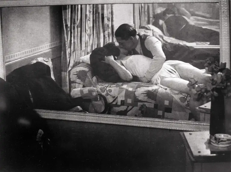 Brassaï black and white vintage photography poster featuring a passionate couple kissing on a patterned bed, reflected in a mirror.