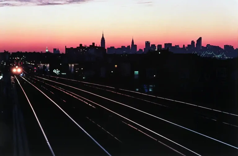 Bruce Davidson photography poster featuring the elevated train tracks of Queens leading toward the dark New York City skyline under a vibrant pink sunset.