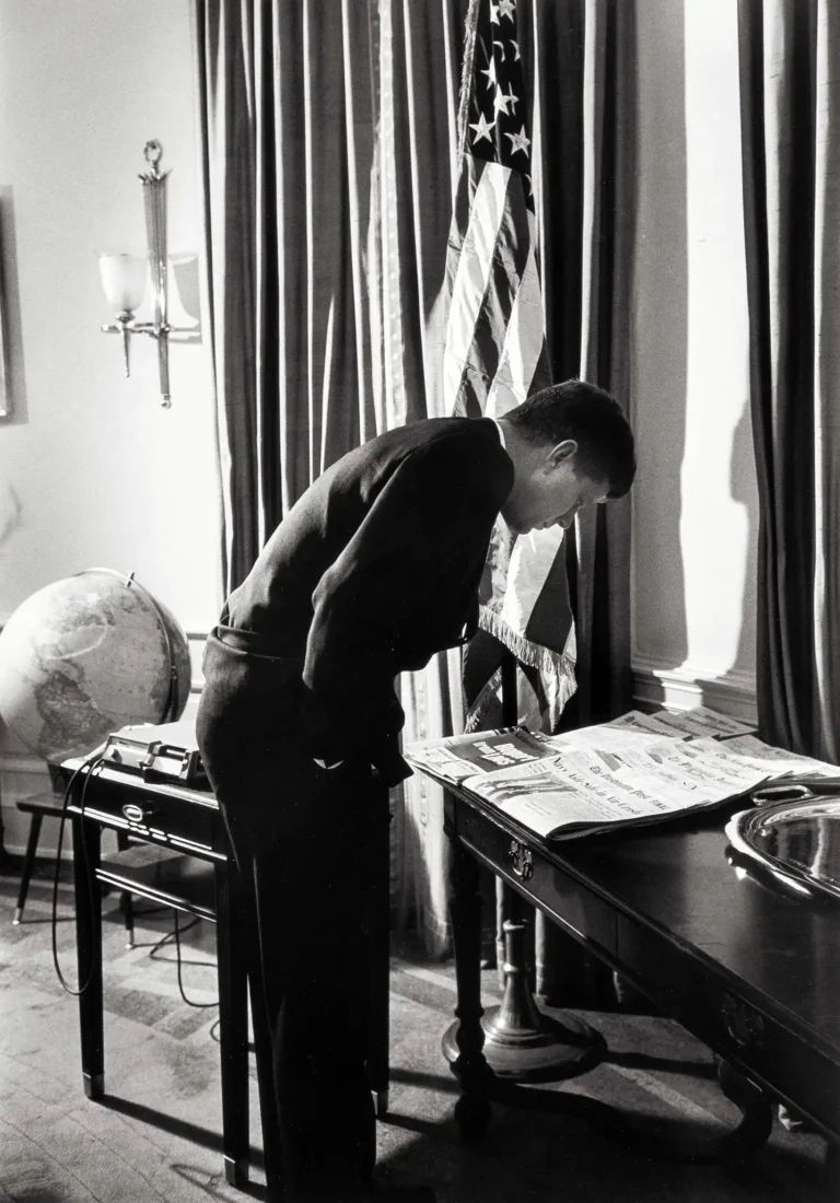 John F. Kennedy historical photo poster, captured by Alfred Eisenstaedt, showing JFK leaning over a desk reading a newspaper in the Oval Office.