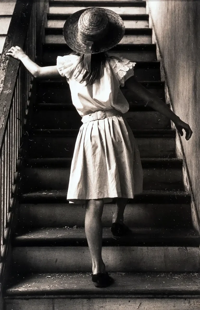 George Krause fine art photography poster featuring a young girl wearing a hat, seen from behind, walking up a dark, moody, wooden staircase.