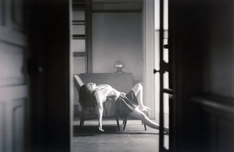 Hisaji Hara Fine Art poster featuring a Japanese schoolgirl reclining dramatically on a chair, viewed through a dark, architectural doorway. High-contrast black and white photograph.