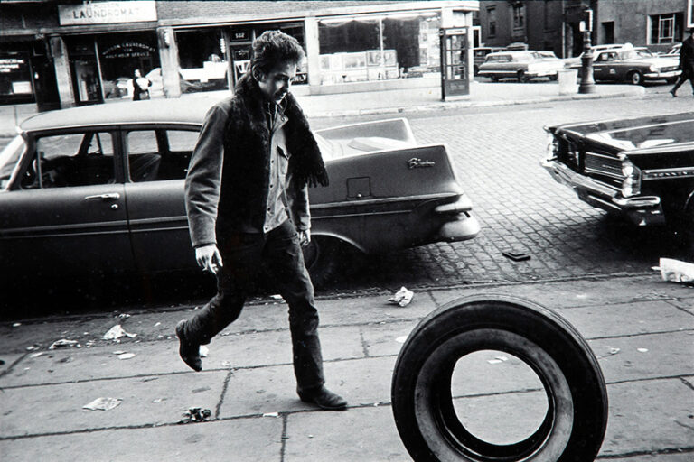 Bob Dylan Music poster photographed by Jim Marshall, featuring Dylan walking on a 1960s cobblestone street in black and white, holding a cigarette.