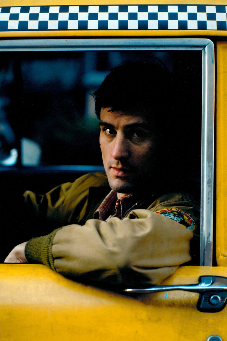 Robert De Niro as Travis Bickle in the classic film Taxi Driver, featuring an intense, direct gaze from the window of his iconic yellow New York City cab.
