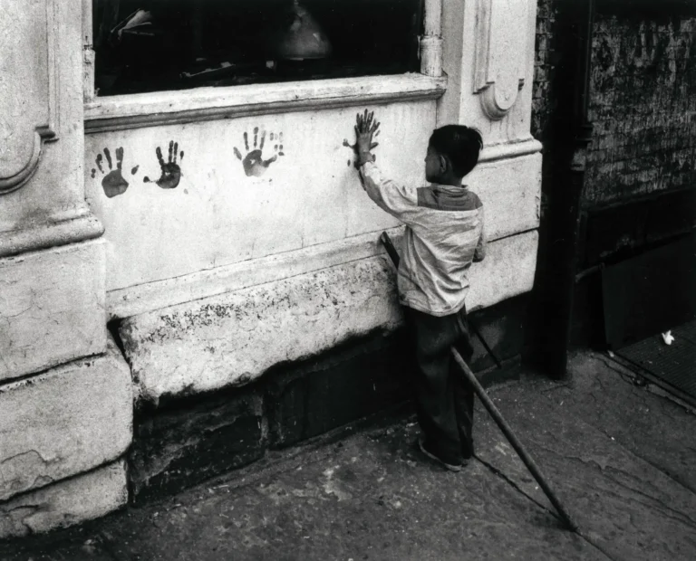 Black and white photography poster showing a young boy reaching up to place a handprint on a grungy, urban wall, capturing an intimate street scene.