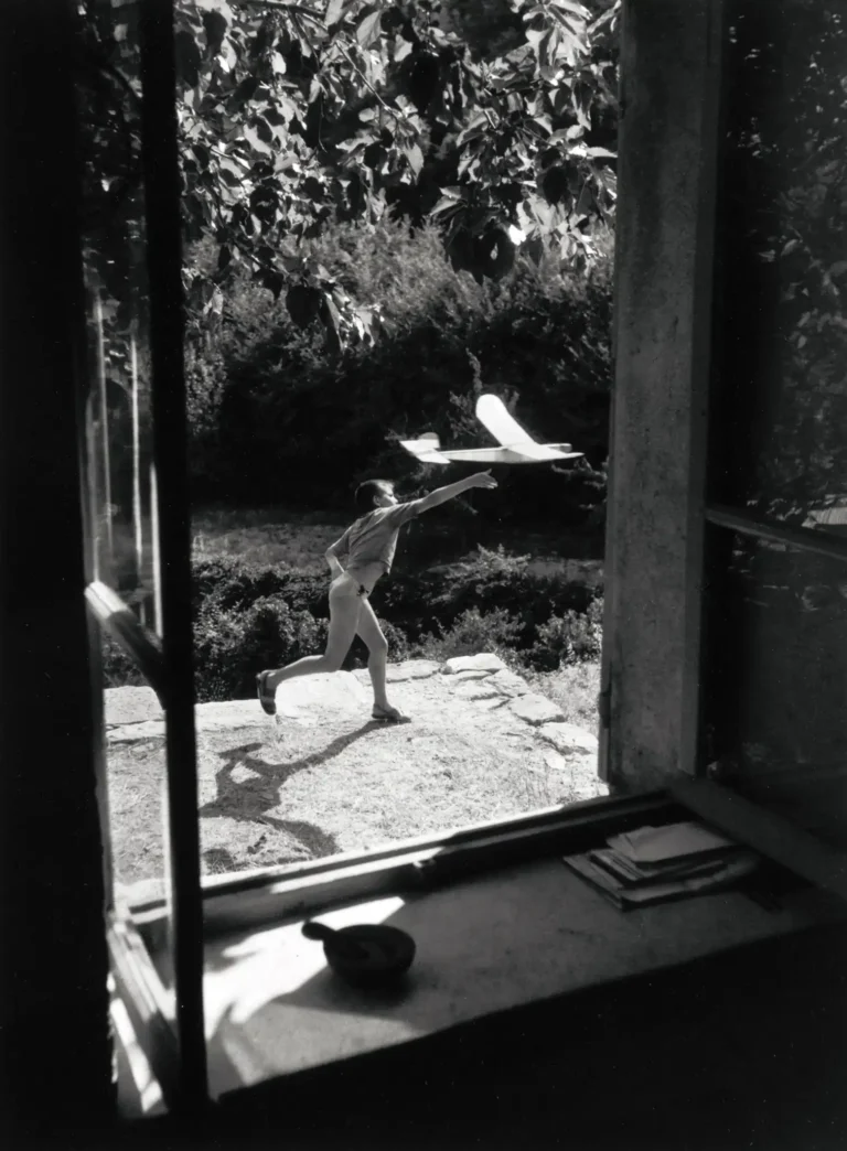 Willy Ronis photography poster featuring a black and white scene of a child running outdoors, framed by a window, launching a toy airplane.