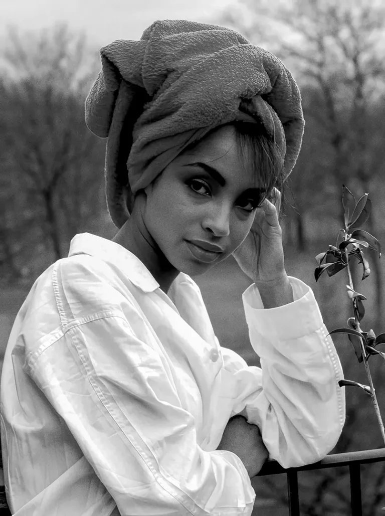 Sade Adu poster in black and white featuring the singer wearing a white shirt and a towel wrap while leaning on a balcony.