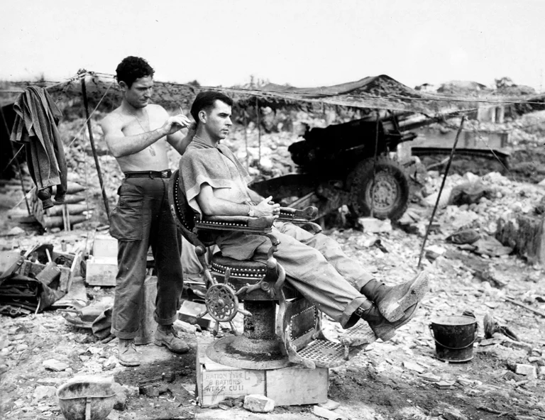 Vintage army barber print showing a soldier receiving a haircut in an ornate chair among WWII rubble and artillery.