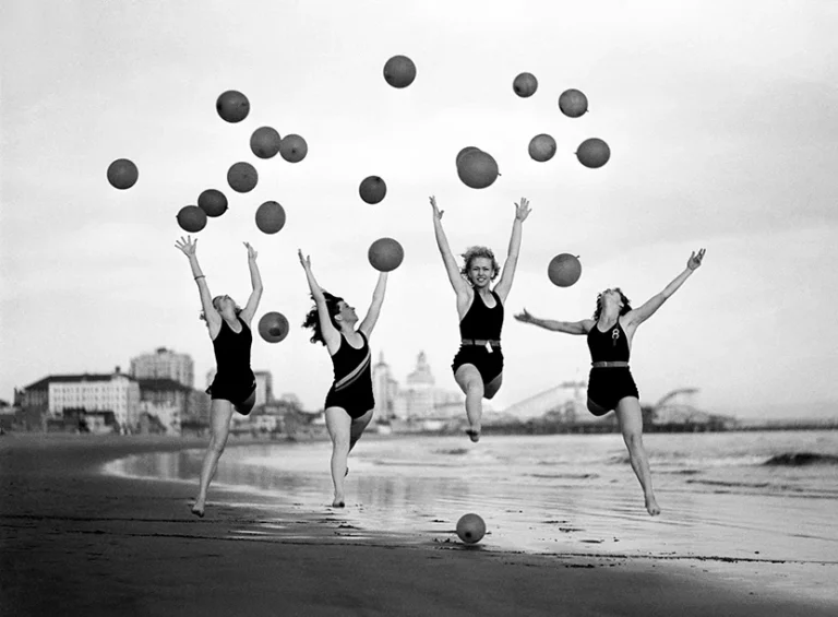 Vintage black and white beach print showing four women jumping on sand with floating balloons near a pier.