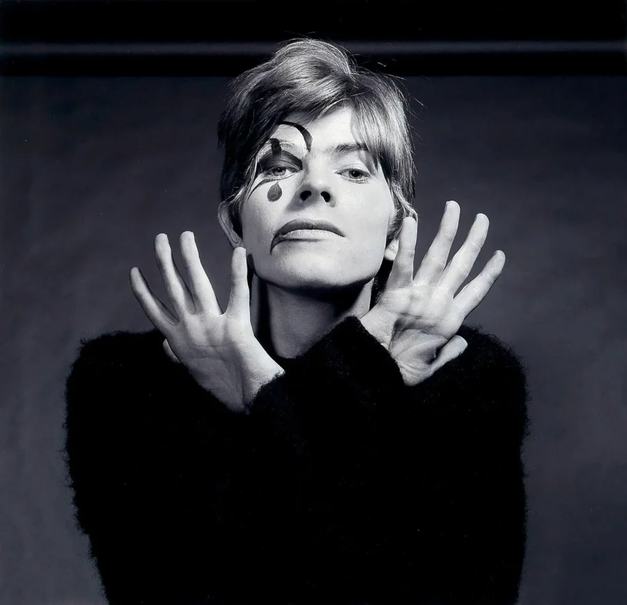 David Bowie in black and white with Aladdin Sane face paint holding his hands up in a theatrical pose.