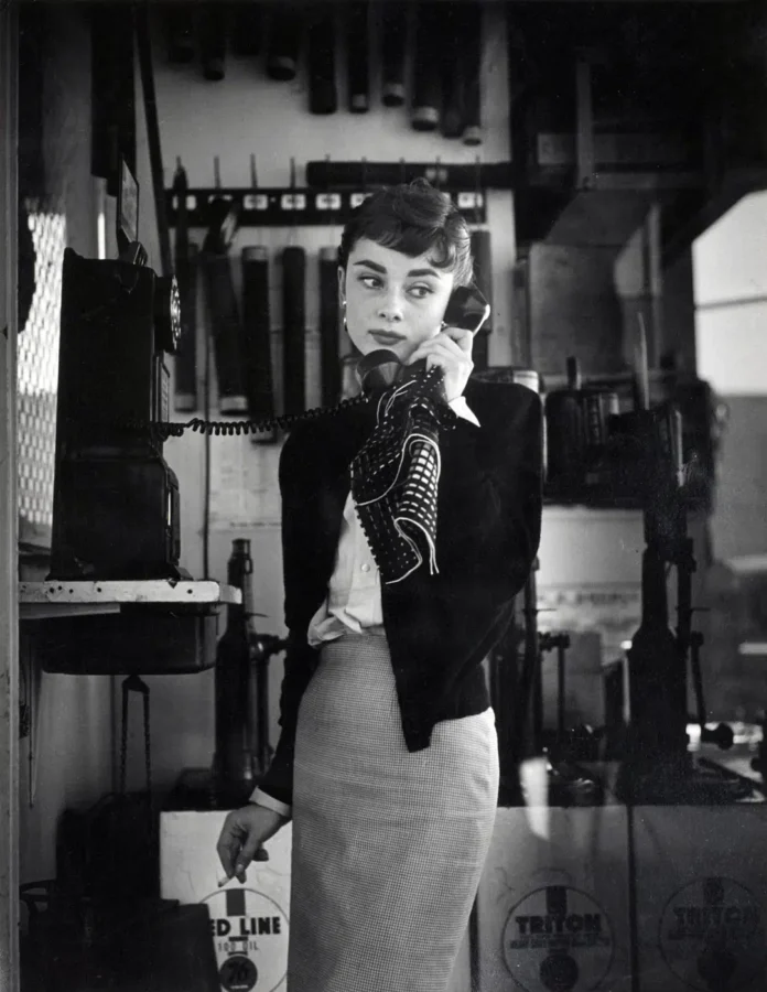 Audrey Hepburn in a black and white photo talking on a payphone while holding a cigarette, wearing a scarf.