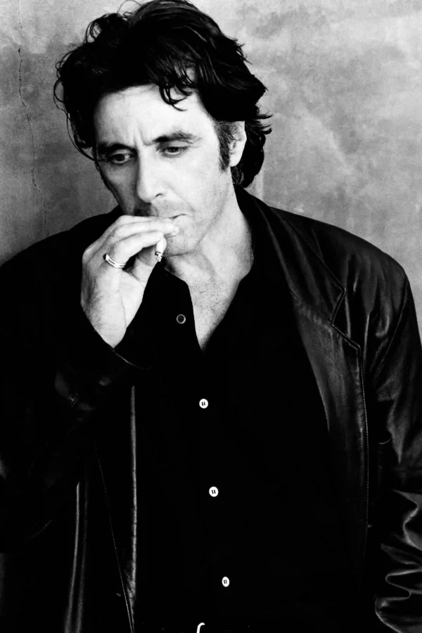 High-contrast black and white photography of Al Pacino wearing a leather jacket and smoking, looking downward with a pensive expression.