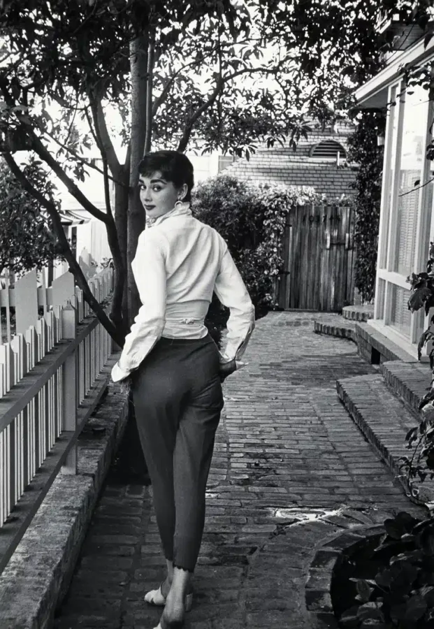 Black and white 1953 photograph of Audrey Hepburn looking over her shoulder on a garden path with a white fence.
