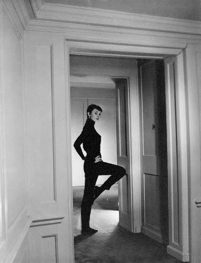 Black and white portrait of Audrey Hepburn in a black turtleneck standing in a paneled hallway in 1955.