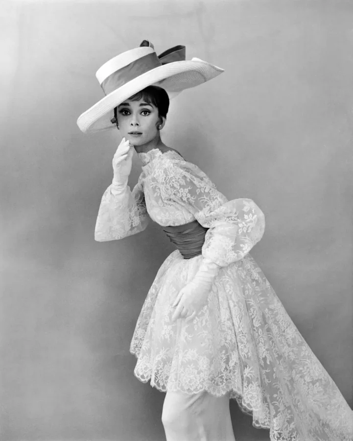 Black and white portrait of Audrey Hepburn wearing an ornate lace dress and wide brim hat in a 1963 fashion photography print.