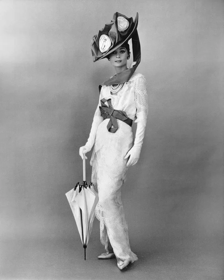 Audrey Hepburn in a white lace dress and large floral hat holding a parasol in a 1963 black and white cinema portrait.