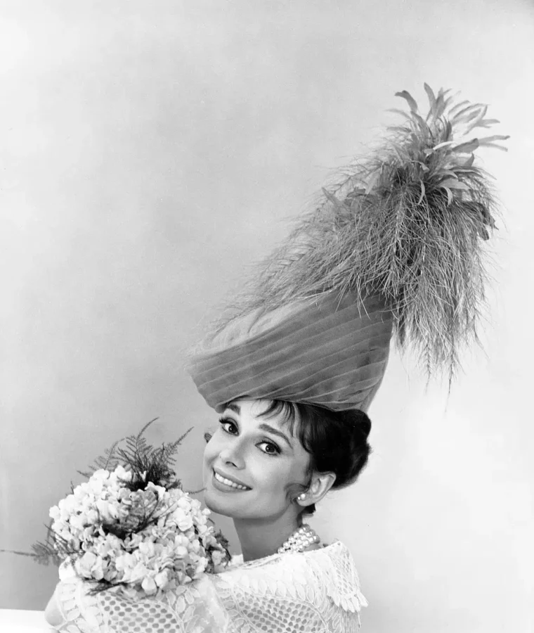 Black and white 1963 portrait of Audrey Hepburn wearing an ornate feathered turban hat and holding a large bouquet of flowers.