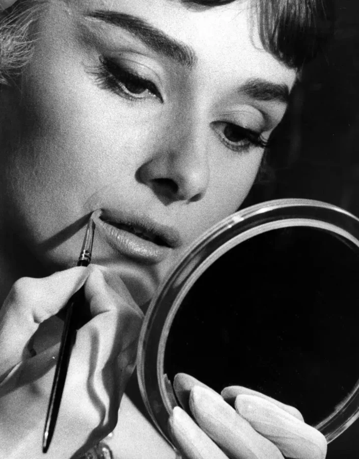 Audrey Hepburn applying lipstick with a brush in a 1957 black and white glamour photography print featuring a circular hand mirror.