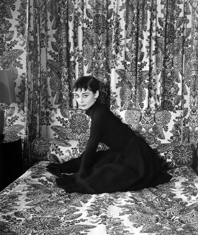 Black and white photograph of Audrey Hepburn in 1954 wearing a black turtleneck and skirt seated on a floral patterned bed.