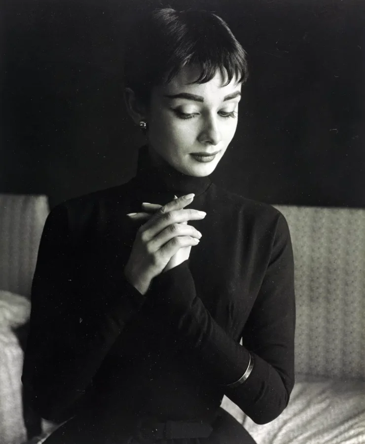 Black and white archival portrait of Audrey Hepburn from March 1954 wearing a dark turtleneck with her hands clasped.