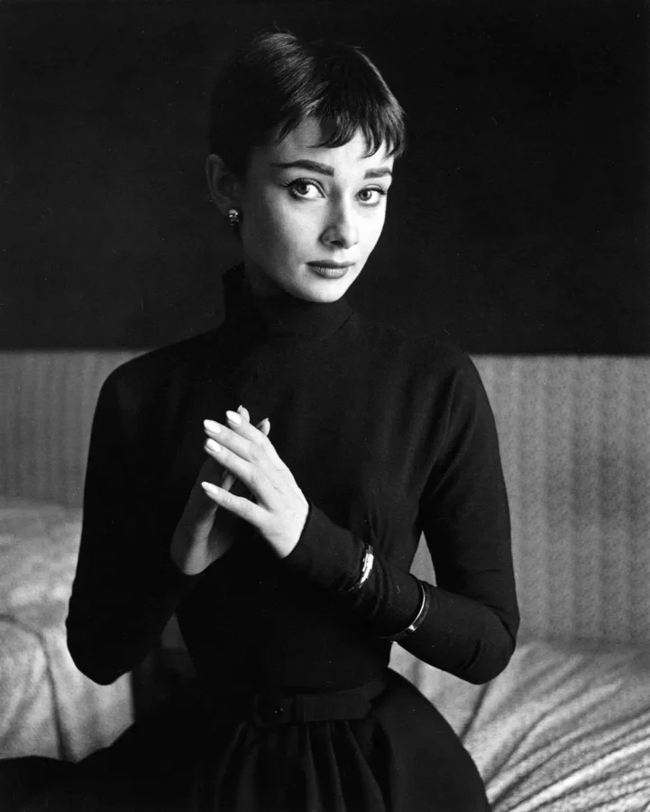 Black and white publicity portrait of Audrey Hepburn in a dark turtleneck with her hands clasped, March 1954.