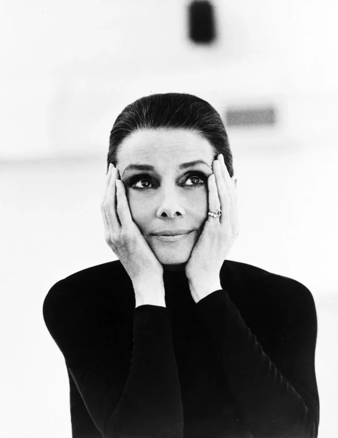 Audrey Hepburn in a high-contrast black and white photography portrait for Vanity Fair, wearing a black turtleneck with hands on her cheeks.