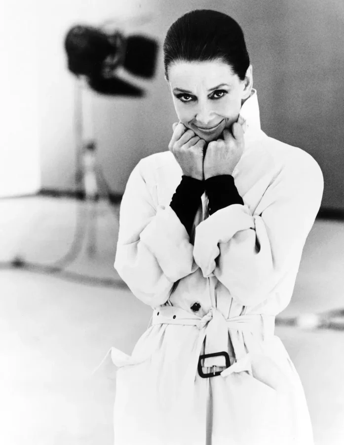 Black and white portrait of Audrey Hepburn in a trench coat from her 1991 Vanity Fair session with hands near her face.
