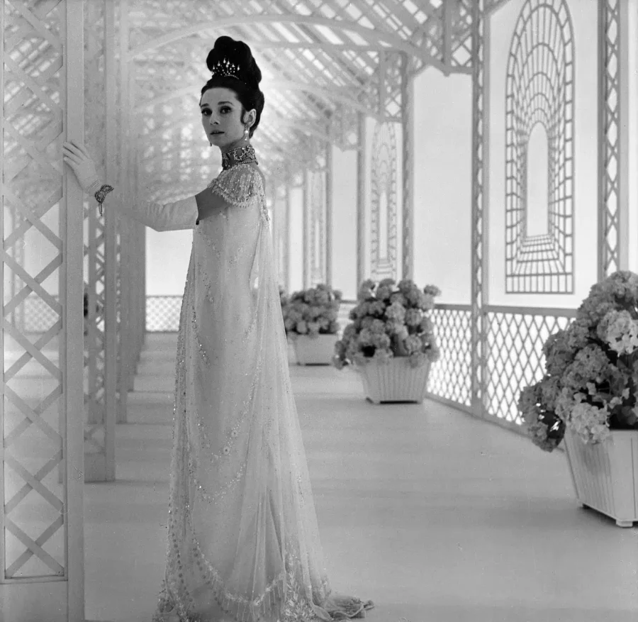 Audrey Hepburn as Eliza Doolittle in a white lace ballgown standing in a conservatory with white trellises and hydrangea flowers.