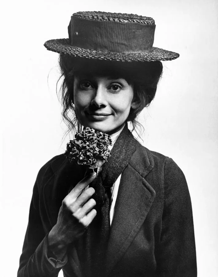Black and white portrait of Audrey Hepburn as Eliza Doolittle in a boater hat holding a small bunch of flowers.