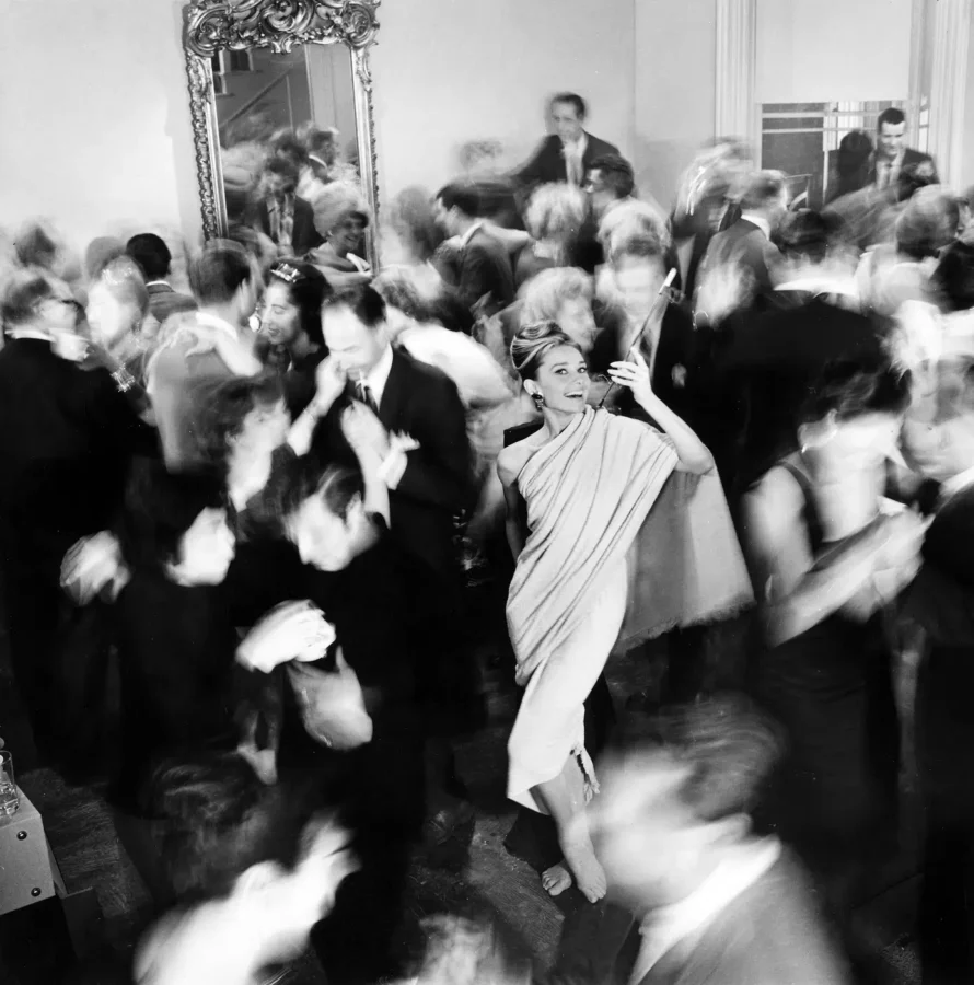 Black and white candid photo of Audrey Hepburn as Holly Golightly barefoot at a party with cigarette holder and blurred guests.
