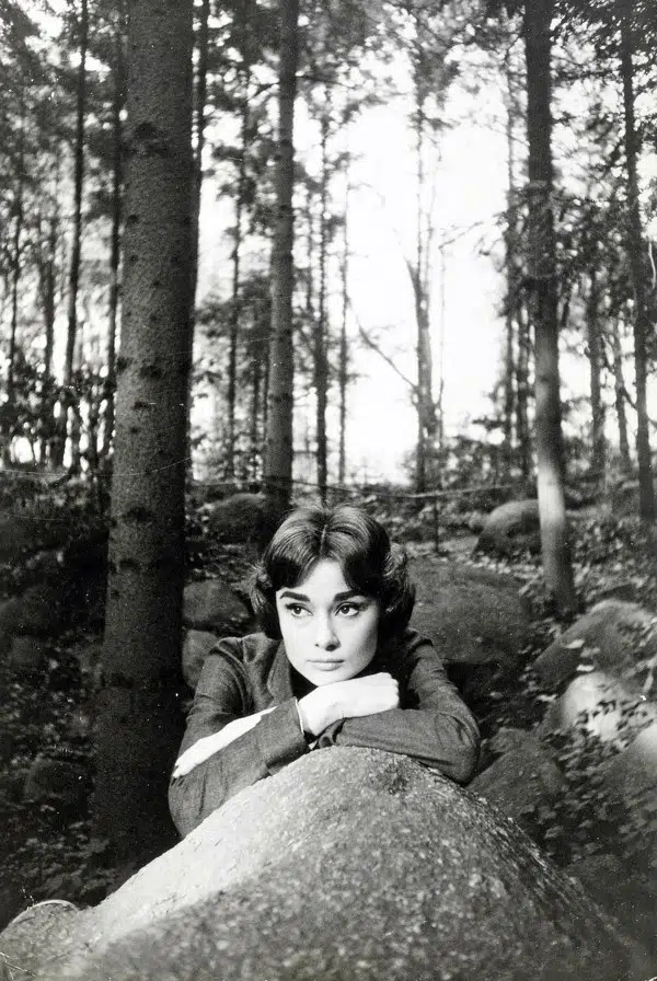 Black and white photograph of Audrey Hepburn leaning on a rock in a Paris forest during the 1957 filming of Love in the Afternoon.