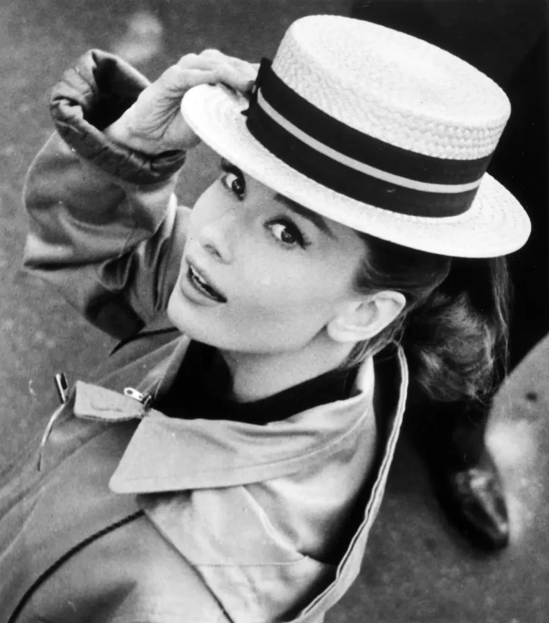 Audrey Hepburn wearing a straw boater hat and trench coat in a black and white 1950s film production photograph.