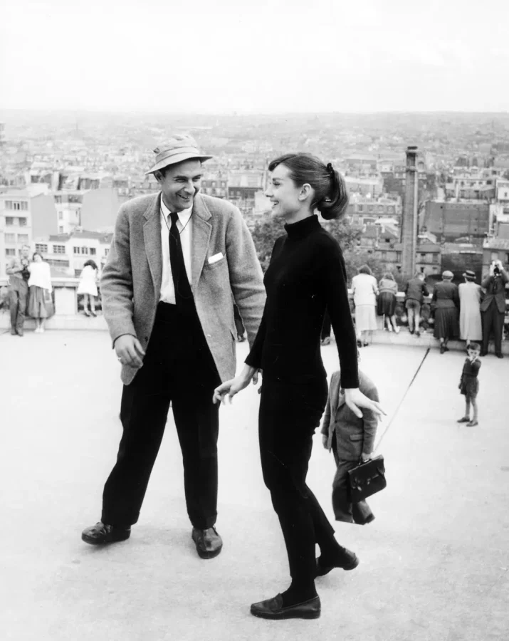 Audrey Hepburn laughing on the Funny Face film set in Paris with a city skyline background in black and white photography.