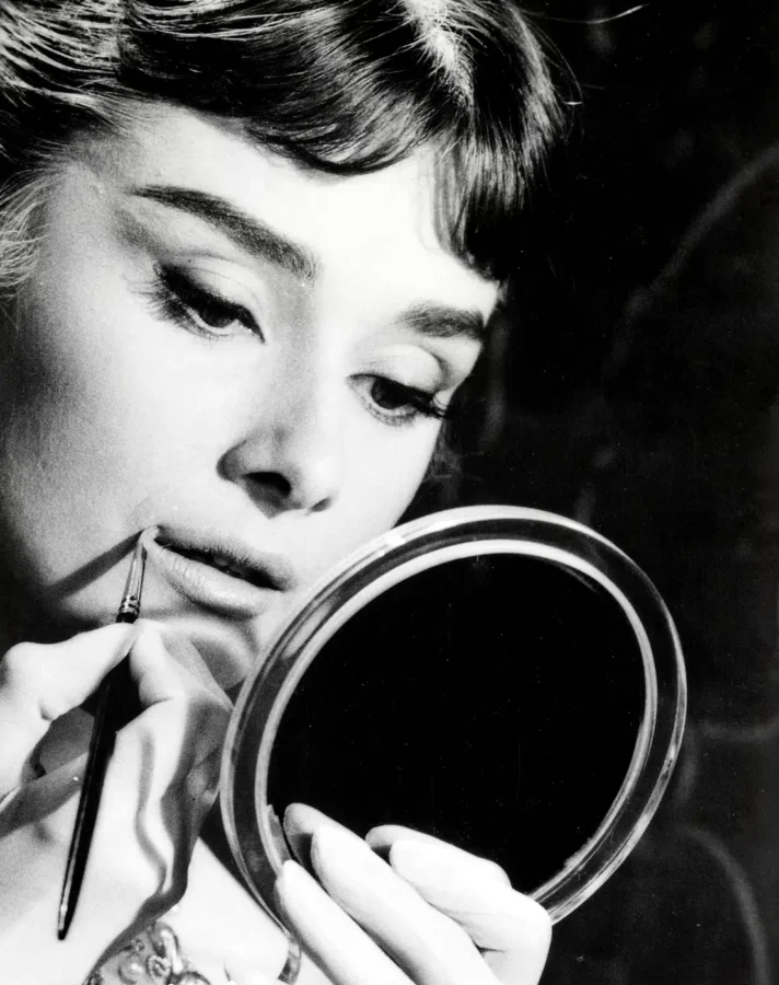 Audrey Hepburn applying lipstick in a round mirror on the Rome set of War and Peace in black and white photography.