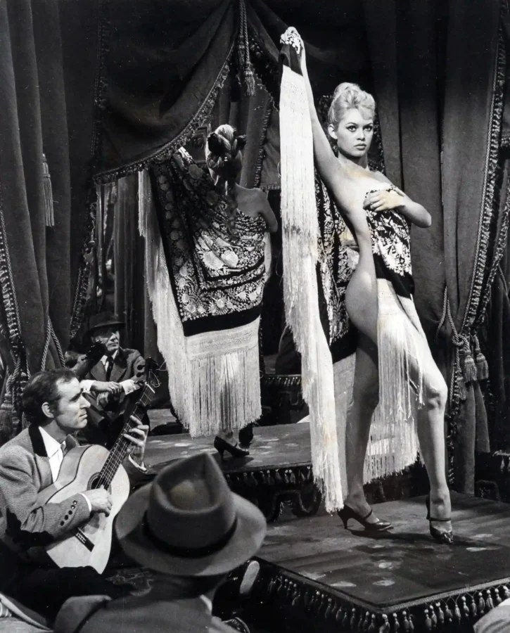 Brigitte Bardot in a fringed stage costume with a floral shawl on a theatrical stage with guitarists in black and white.