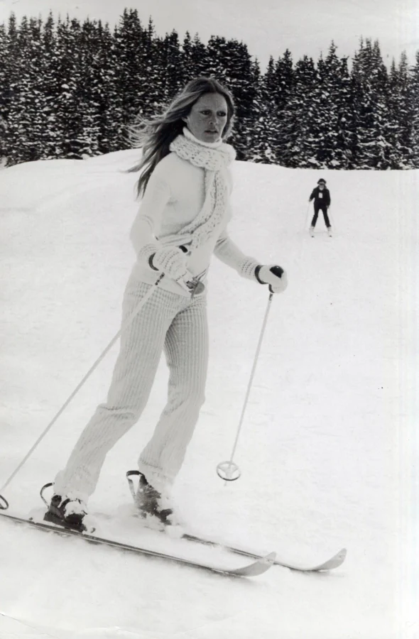 Black and white photo of Brigitte Bardot skiing in Switzerland wearing a white knit scarf and winter outfit against a snowy forest.