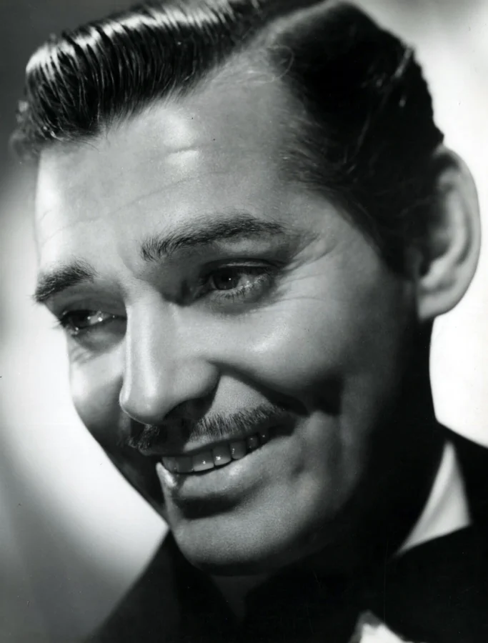 Black and white close-up portrait of Clark Gable smiling with his signature moustache and slicked hair in high-contrast studio lighting.