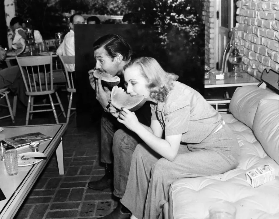 Black and white candid photo of Clark Gable and Carole Lombard eating watermelon slices on a patio bench in 1939.