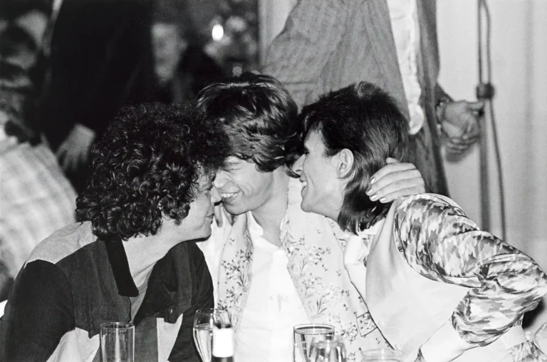 Clean artwork view of David Bowie, Mick Jagger and Lou Reed Cafe Royal Photography Print. Candid black and white photograph of David Bowie, Mick Jagger, and Lou Reed sharing an intimate moment at a table during a 1973 London after-party.