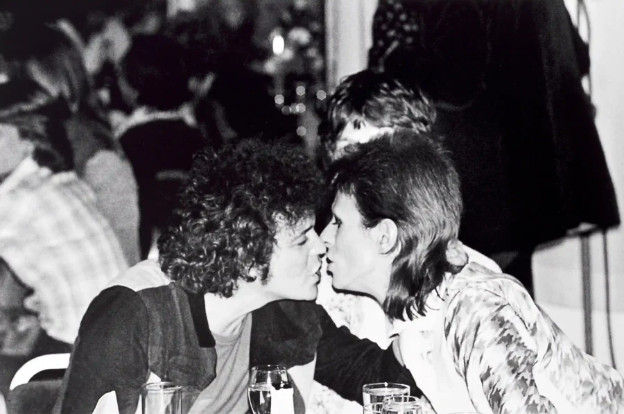 Black and white photograph of David Bowie and Lou Reed leaning in for a kiss at a 1973 party with high contrast and film grain.