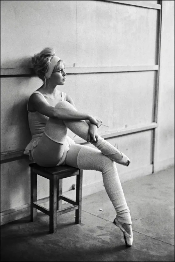 Brigitte Bardot in a dance studio wearing a leotard and leg warmers sitting on a stool in a black and white photograph.