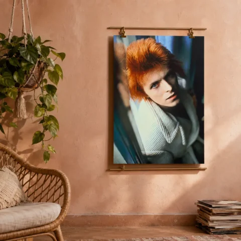 A framed ziggy stardust art print with vibrant orange hair hangs on a modern gray wall above a minimalist wooden desk.