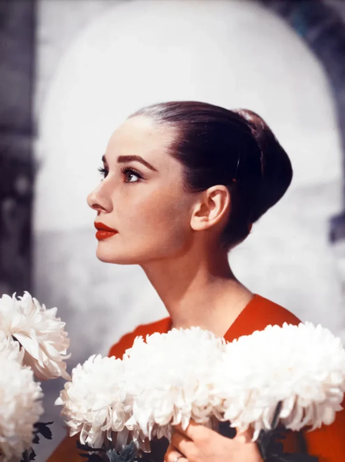 Side profile of Audrey Hepburn in a red dress holding white flowers with bold red lipstick and a sleek hair bun.