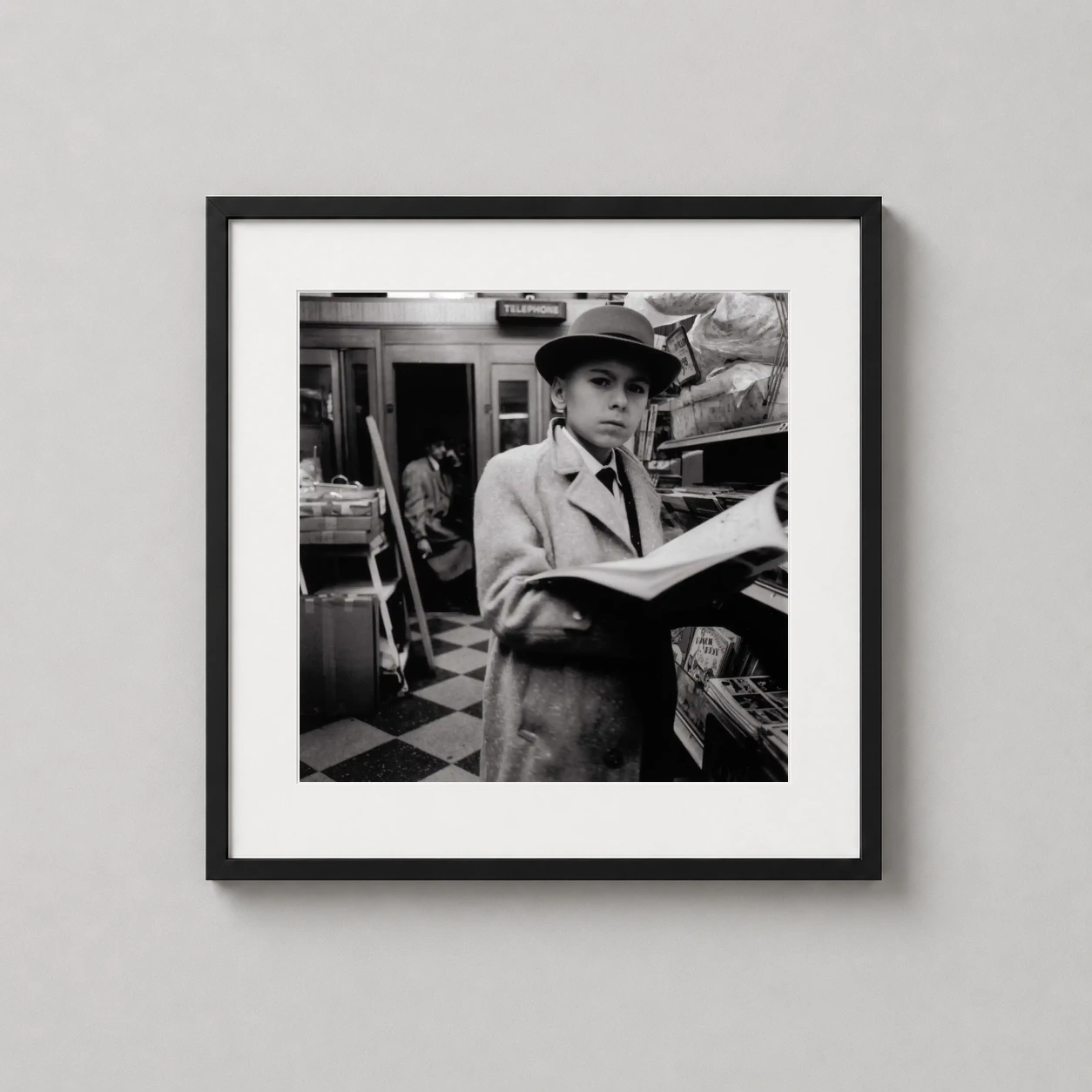 Room mockup of Diane Arbus Boy in Fedora Reading a Magazine, NYC 1956 Photography Print, displayed as framed wall art in a office setting as a square print with black and white tones.