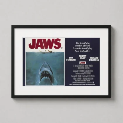 A landscape Jaws horror movie poster displayed in a black frame on a neutral wall in a modern living room setting.