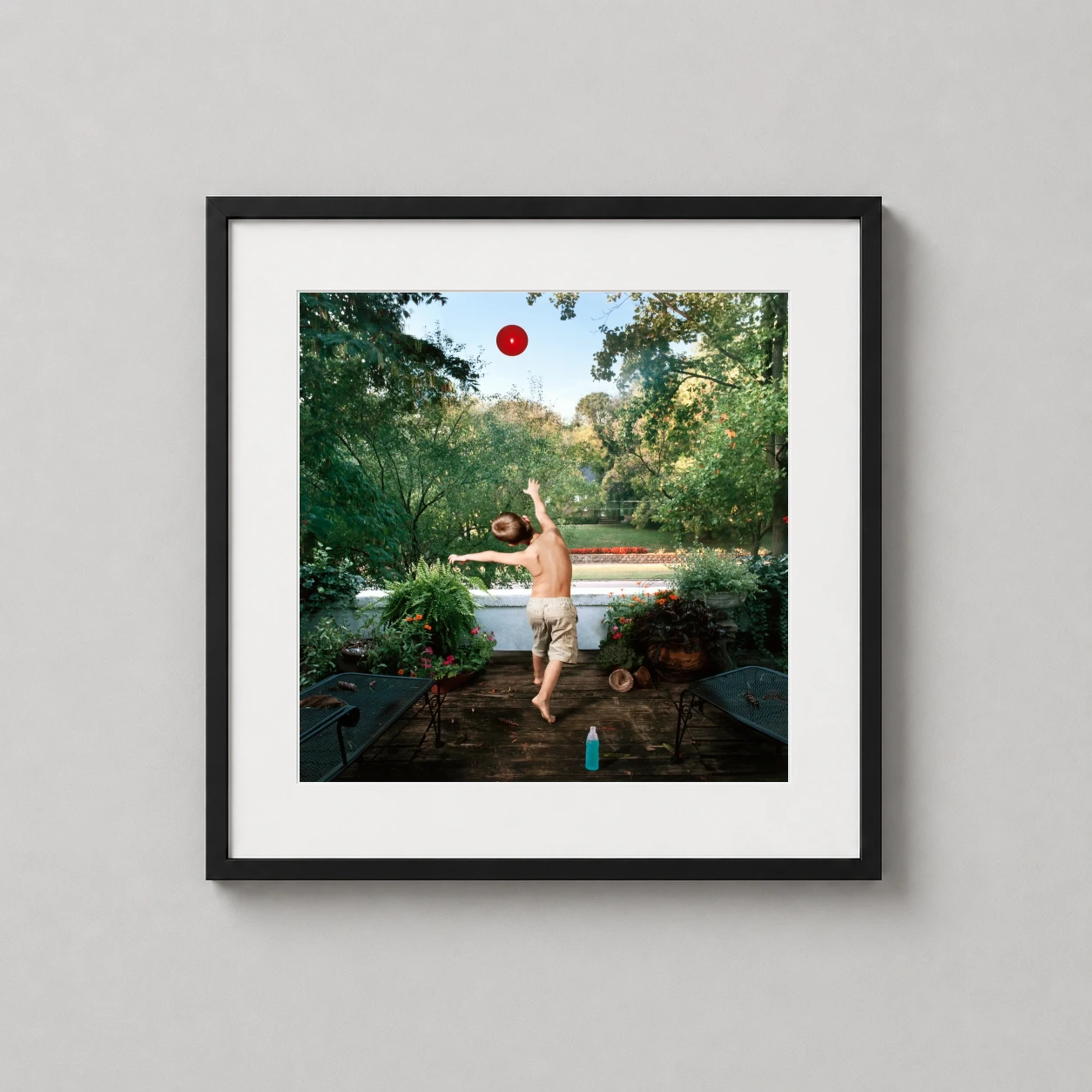 Julie Blackmon Powerade — young boy reaches for a suspended red ball on a lush garden deck in