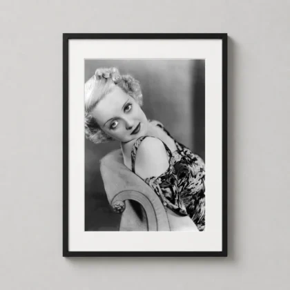A framed Bette Davis portrait print hangs on a neutral wall above a vintage wooden side table in a well-lit room.