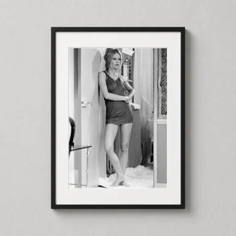 A vertical framed art print of Brigitte Bardot in Les Novices displayed on a neutral bedroom wall near a white lamp.