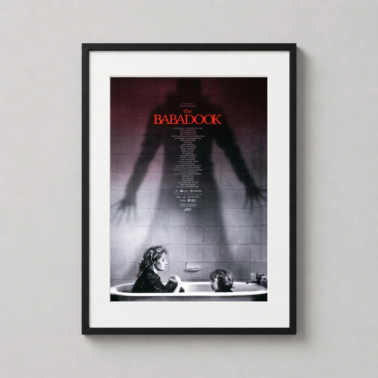 Room mockup of The Babadook Horror Movie Poster by Greg Ruth Mondo Poster, displayed as framed wall art in a home theater setting as a portrait print with black and white and red tones.
